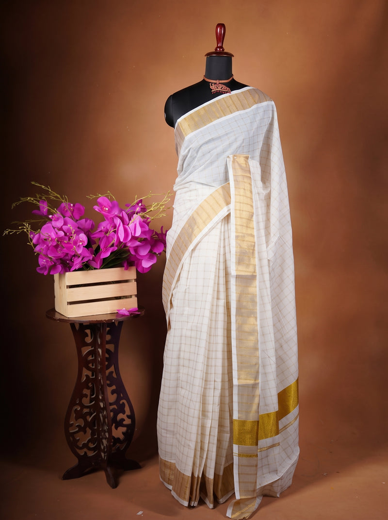 A white and gold saree draped on a black mannequin, styled next to a wooden crate filled with bright pink flowers on a decorative dark stand, set against a warm brown studio backdrop.
