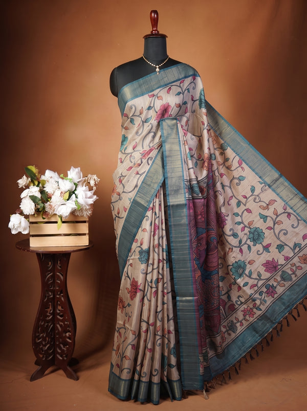 A patterned saree displayed on a black mannequin against a warm brown studio backdrop, with a decorative wooden stand holding white flowers at the left. The saree has a beige base with blue, purple, and pink floral motifs and a blue border.