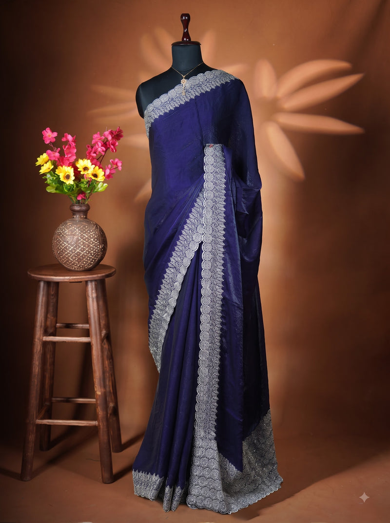 A dark blue saree with intricate silver border embroidery draped on a black mannequin, styled next to a wooden stool holding a vase of colorful flowers against a warm brown backdrop.