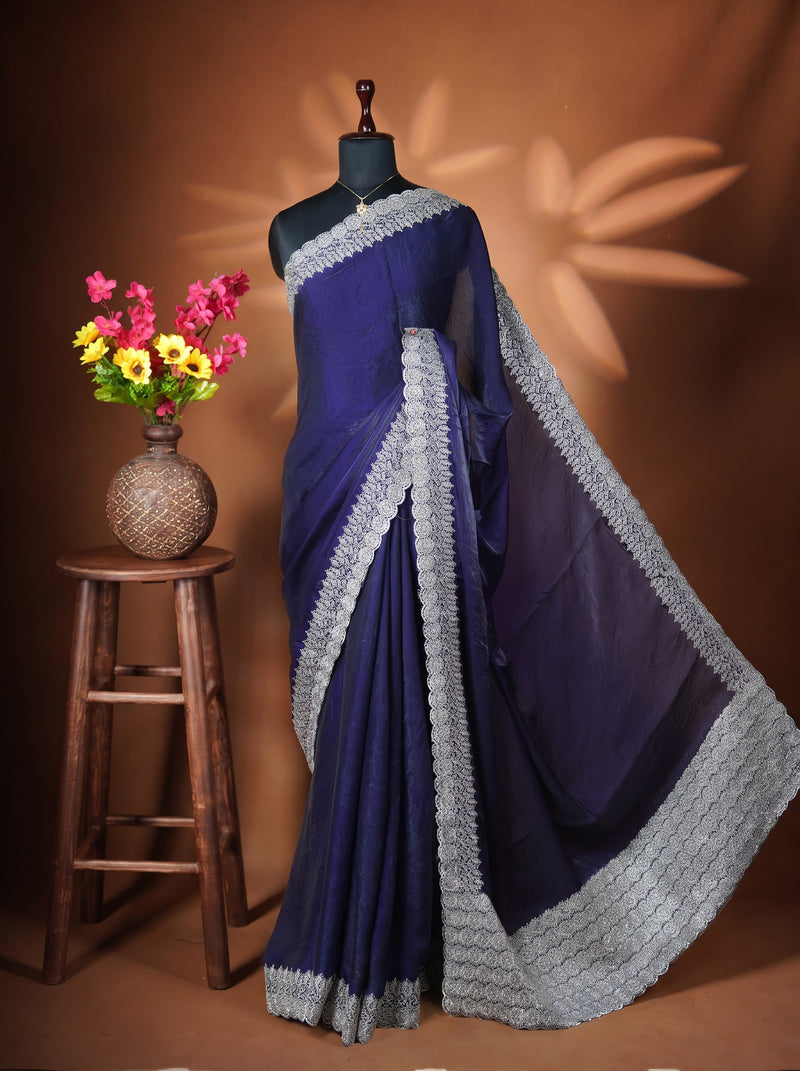 A dark blue-purple saree with a wide light gray embroidered border is draped on a black mannequin torso. The mannequin is set against a warm brown studio backdrop with leaf-shaped shadows, and a small wooden stool with a vase of colorful flowers is visible on the left.