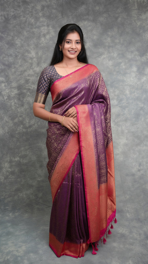 A woman modeling a purple and pink saree with gold zari patterns, standing against a textured gray backdrop.