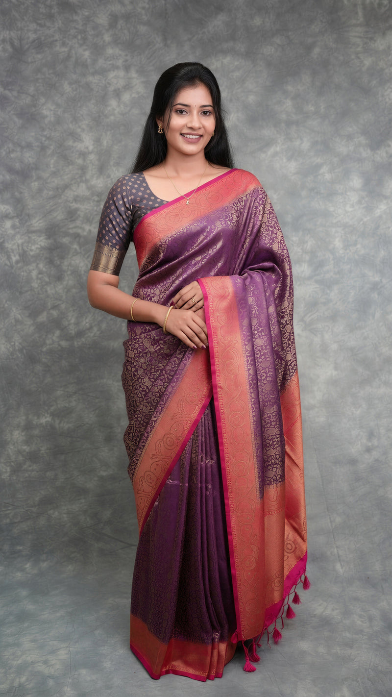 A woman modeling a purple and pink saree with gold zari patterns, standing against a textured gray backdrop.