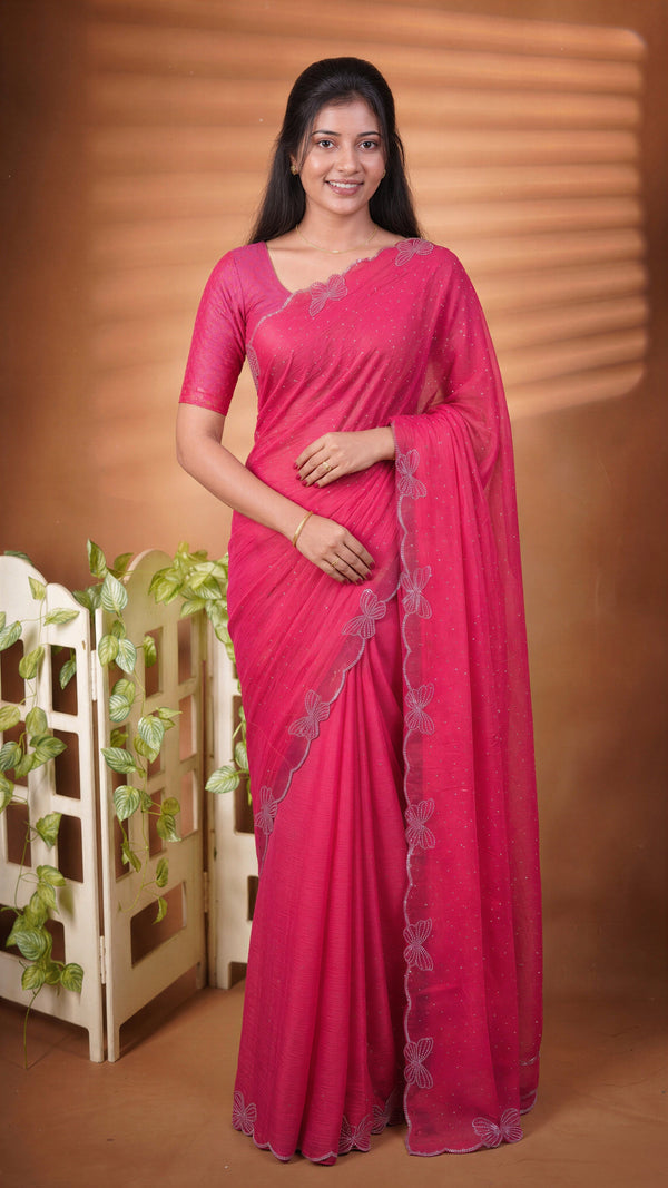 A woman modeling a bright pink saree with a matching blouse, standing in front of a warm brown backdrop with decorative plants on the left.