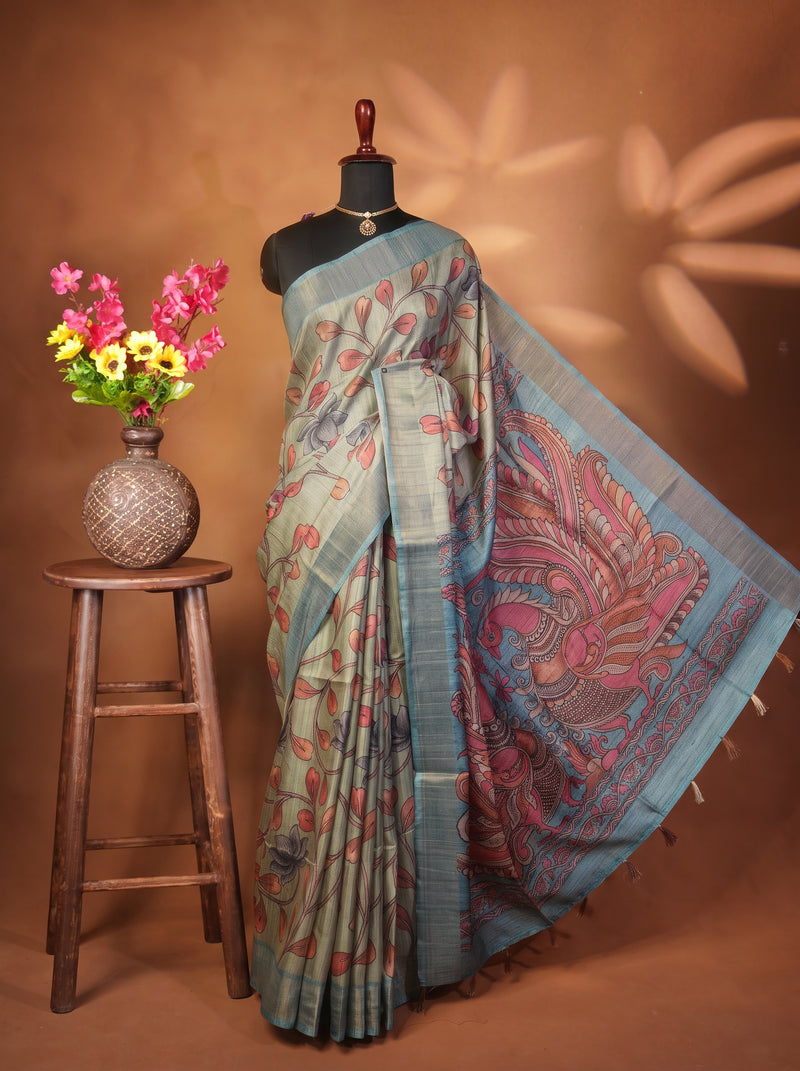 A light blue and pink printed saree displayed on a black mannequin torso, styled with a decorative brown vase of flowers on a wooden stool against a warm brown backdrop.