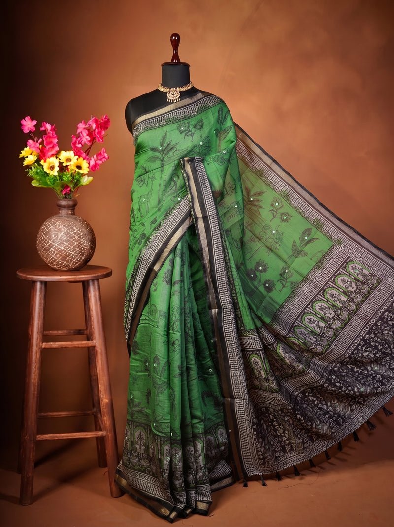 A green saree with black and silver border patterns displayed on a black mannequin, set against a warm brown studio backdrop. A small wooden stool on the left holds a woven vase filled with pink, yellow, and red flowers.