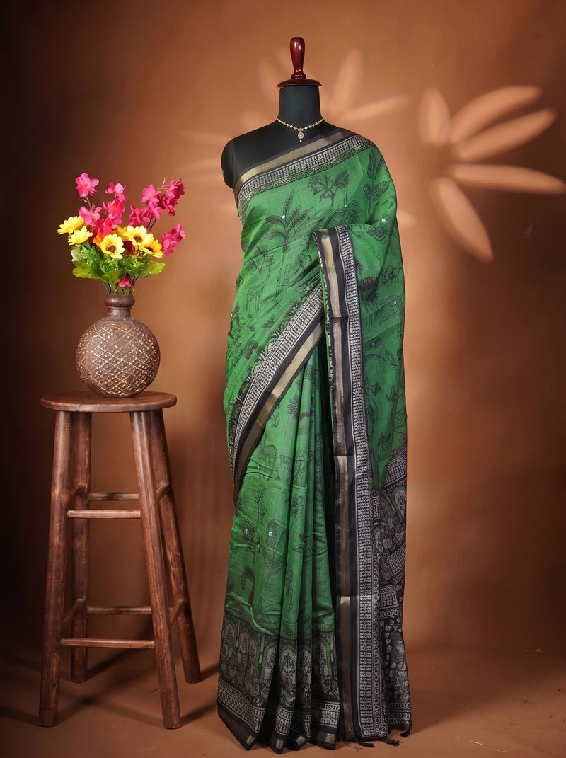 A green silk saree with black and silver patterns and borders, draped on a black mannequin. The mannequin is set against a warm brown backdrop with decorative leaf shadows, and a wooden stool holding a vase of colorful flowers is on the left.