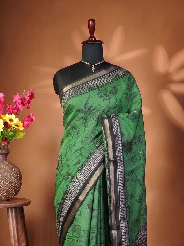 A green silk saree with black and silver patterns and borders, draped on a black mannequin. The mannequin is set against a warm brown backdrop with decorative leaf shadows, and a wooden stool holding a vase of colorful flowers is on the left.