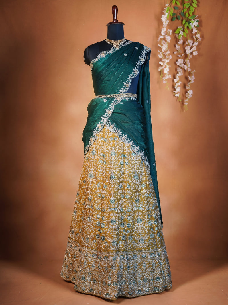 A traditional Indian lehenga choli dress displayed on a black mannequin against a warm brown backdrop. The garment features a deep teal green upper section and drape, paired with a heavily embroidered lower skirt in gold, blue, and white.