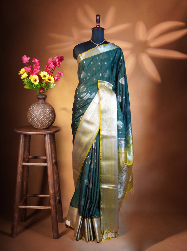 A dark teal green saree with a wide gold zari border and small gold motifs, displayed on a black mannequin. The mannequin is set against a warm brown backdrop with decorative leaf shadows, and a wooden stool with a vase of colorful flowers is positioned to the left.