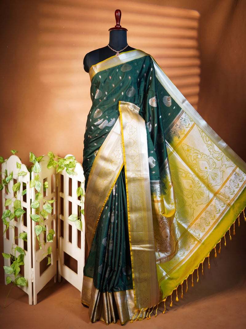 A dark green silk saree with metallic gold and silver zari motifs, draped on a black mannequin. The saree features a wide gold border and a yellow-gold pallu with intricate silver and gold floral patterns.