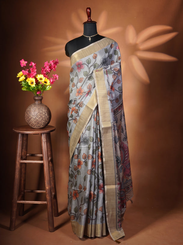 A patterned grey and beige saree draped on a black mannequin torso, styled with a gold necklace. To the left, a wooden stool holds a woven vase filled with pink and yellow flowers against a warm brown backdrop with leaf-shaped shadows.