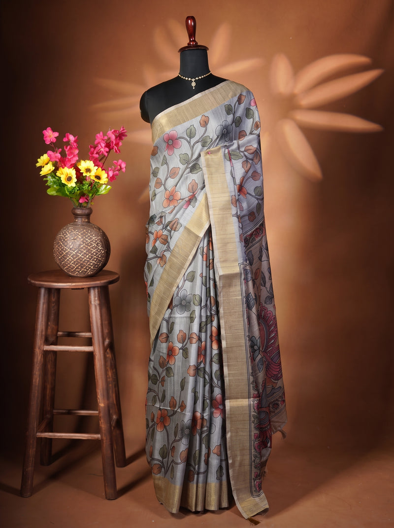 A patterned grey and beige saree draped on a black mannequin torso, styled with a gold necklace. To the left, a wooden stool holds a woven vase filled with pink and yellow flowers against a warm brown backdrop with leaf-shaped shadows.