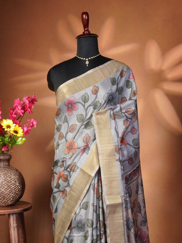 A patterned grey and beige saree draped on a black mannequin torso, styled with a gold necklace. To the left, a wooden stool holds a woven vase filled with pink and yellow flowers against a warm brown backdrop with leaf-shaped shadows.