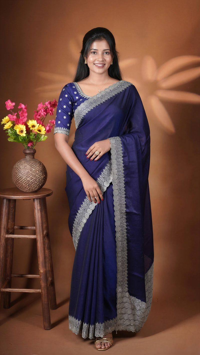 A woman modeling a deep blue saree with a silver embroidered border and a matching blue blouse with white polka dots, standing in front of a warm brown backdrop with a wooden stool and flowers on the left.