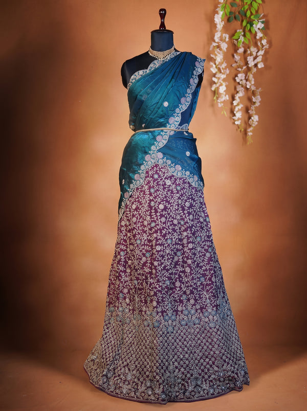 A traditional Indian saree displayed on a black mannequin dress form against a warm brown studio backdrop. The saree features a teal blue upper drape with silver embroidery, a matching embroidered blouse, and a long skirt that transitions from teal to purple with dense silver floral embroidery and a scalloped border.