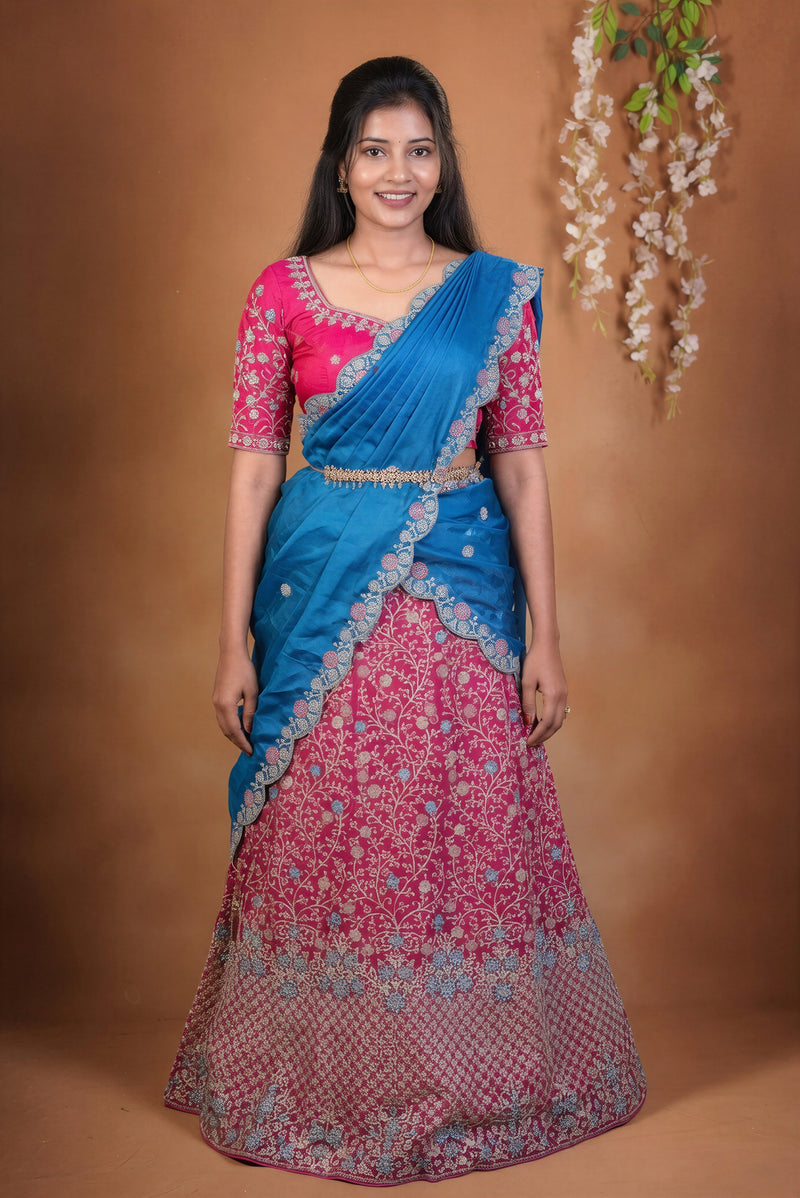A woman modeling a traditional Indian saree with a bright blue upper drape and a pink flared skirt heavily embroidered with floral patterns and gold detailing, set against a warm brown studio backdrop.