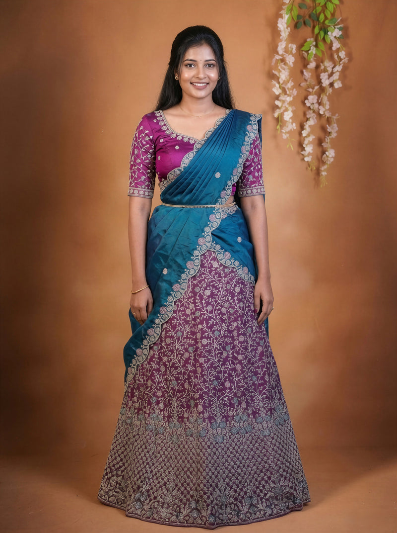 A woman modeling a traditional Indian lehenga choli saree. The saree features a teal blue upper drape and a magenta-purple lower skirt with intricate silver floral embroidery, paired with a matching magenta embroidered blouse.