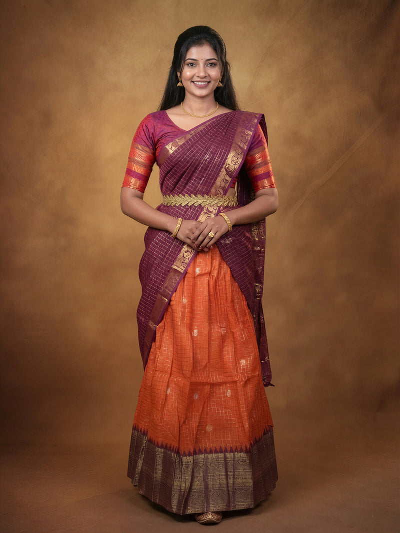 A woman modeling a traditional Indian saree with a deep purple border and a bright orange body, featuring gold zari embroidery and a gold waist belt, set against a warm brown studio backdrop.