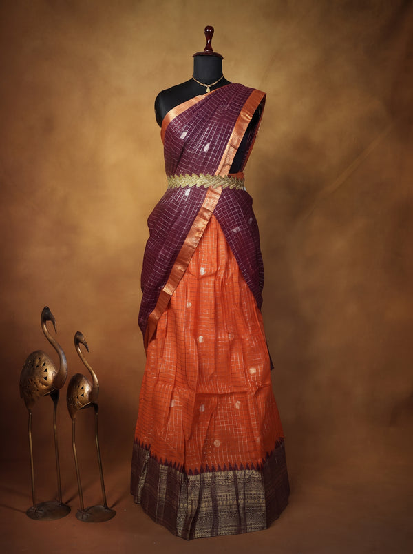 A traditional Indian saree displayed on a black dress form mannequin against a warm brown studio backdrop. The saree has a deep purple upper drape with a gold border, a bright orange body, and a dark patterned border.