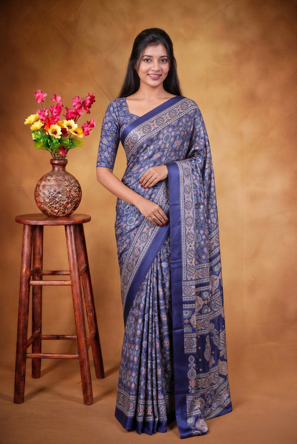 A woman modeling a blue patterned saree with a matching blouse, standing next to a wooden stool with a vase of flowers against a warm brown studio backdrop.