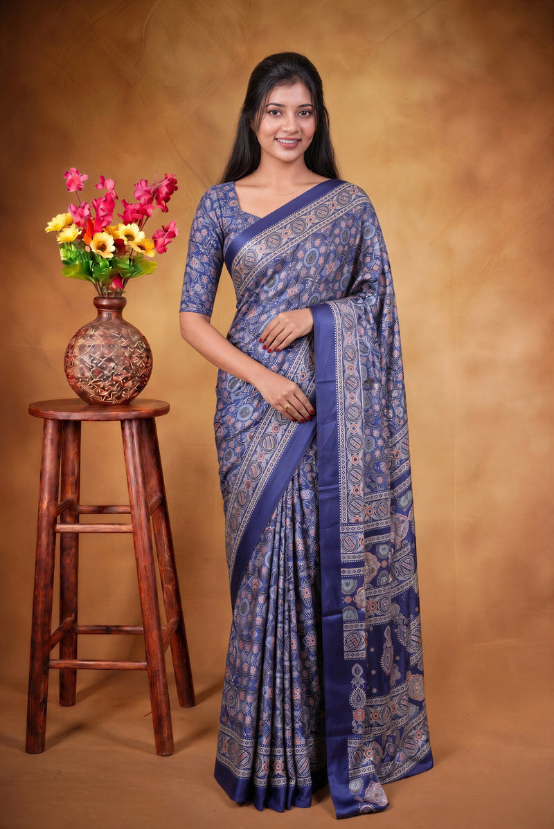 A woman modeling a blue patterned saree with a matching blouse, standing next to a wooden stool with a vase of flowers against a warm brown studio backdrop.