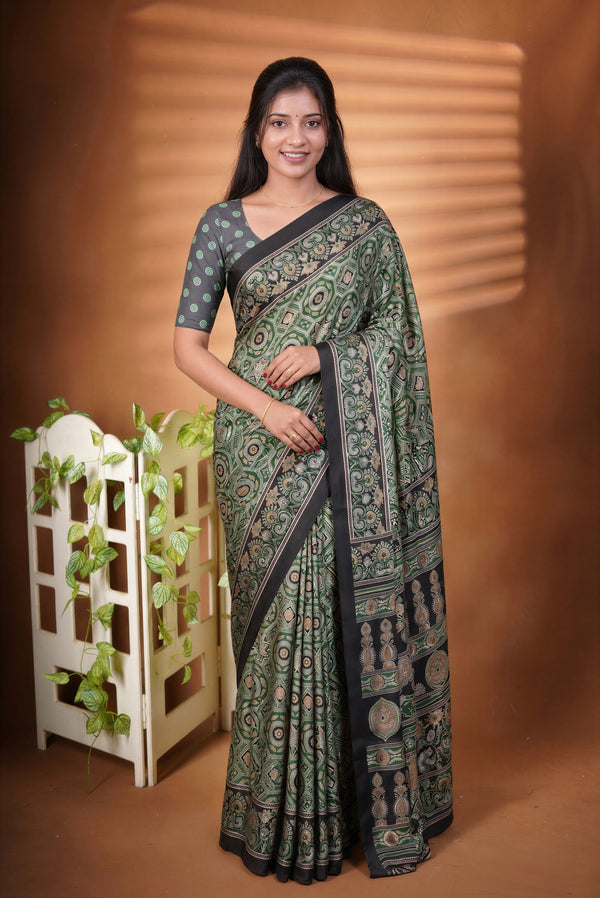 A woman modeling a dark green and black printed saree with a matching printed blouse, standing against a warm brown studio backdrop with a decorative white trellis and green vines on the left.