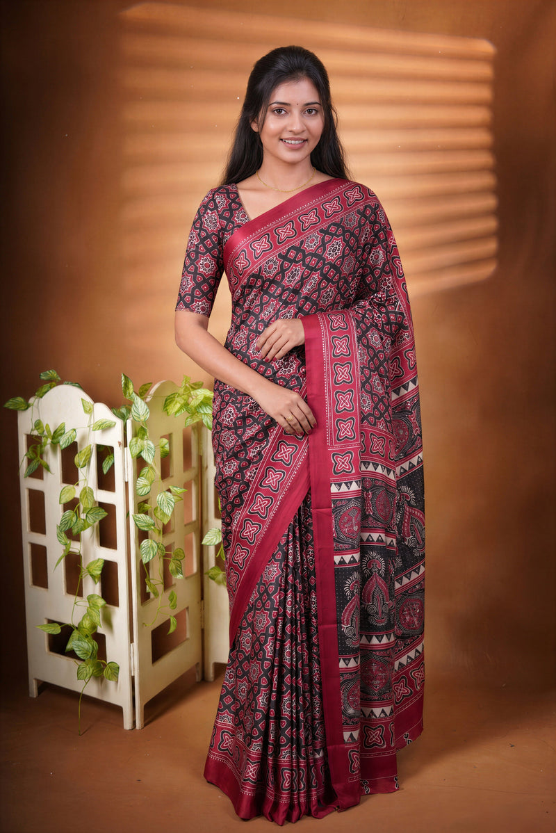 A woman modeling a dark red and black patterned saree with a matching printed blouse, standing against a warm brown studio backdrop with decorative plants on the left.
