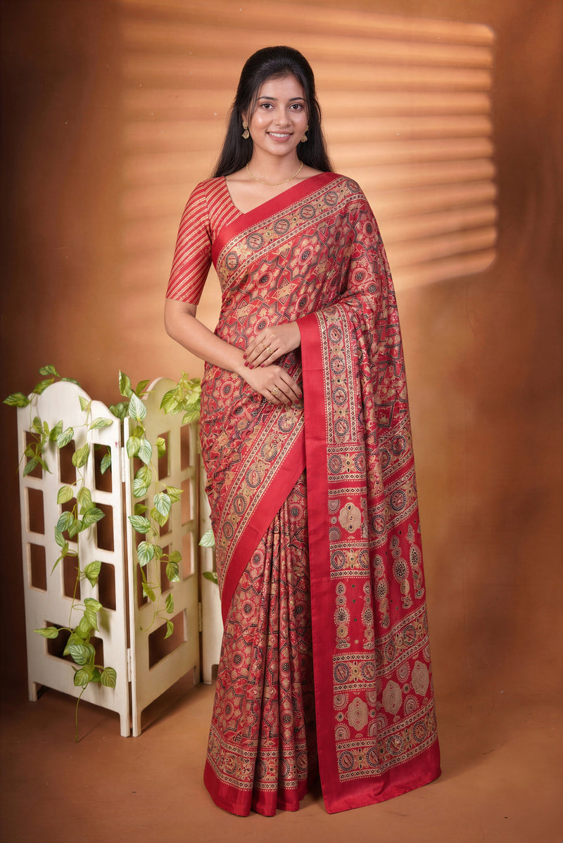 A woman modeling a red and gold patterned saree with a matching red blouse, standing against a warm brown backdrop with decorative plants and a white screen.