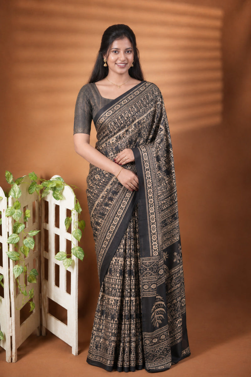 A woman modeling a black and gold patterned saree with a matching black blouse, standing in front of a warm brown backdrop with a small decorative white screen and green leaves on the left.