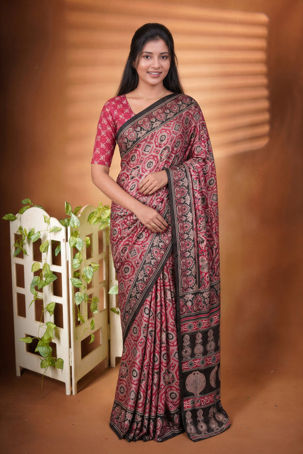 A woman modeling a pink and black patterned saree with a matching printed blouse, standing against a warm brown backdrop with a decorative white screen and green vines on the left.