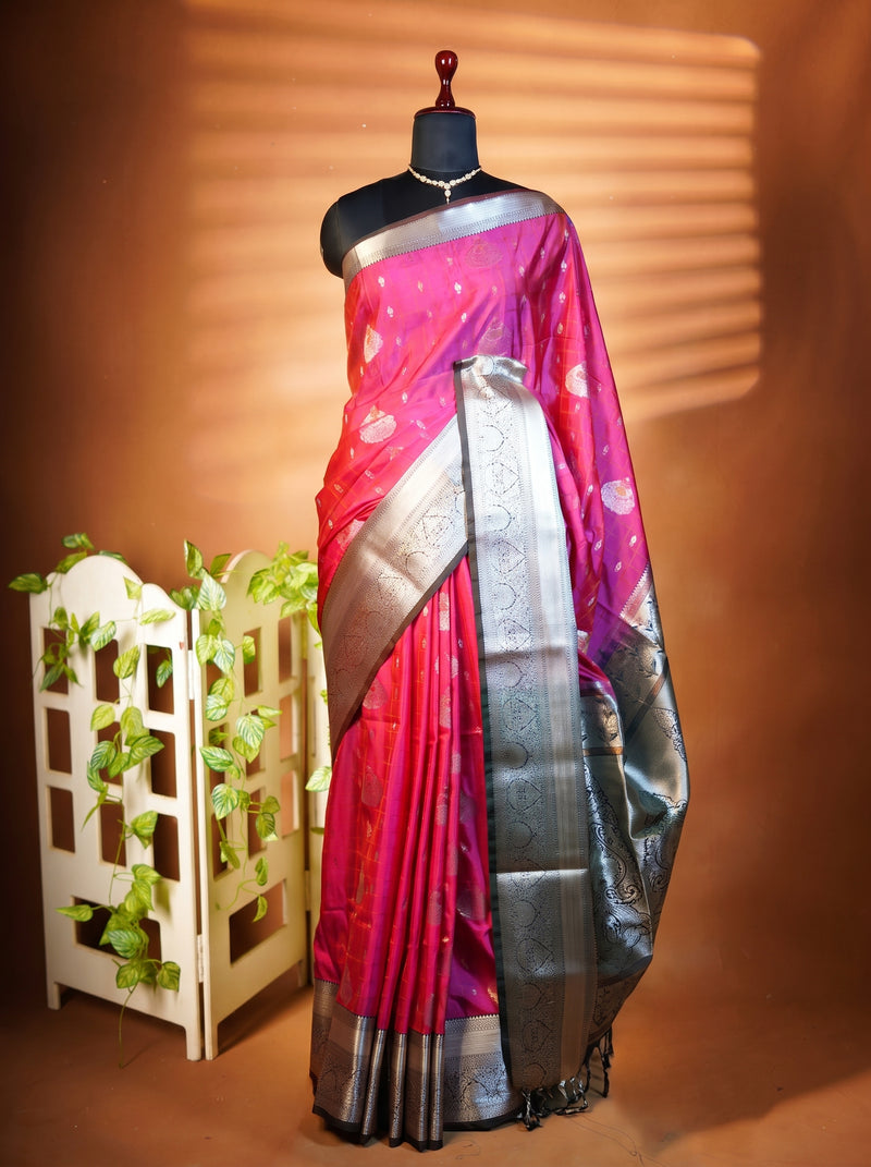 A pink and magenta silk saree with a wide silver metallic border, displayed on a black mannequin torso. The mannequin is set against a warm brown background with decorative shadow patterns, and a small white planter with green vines sits to the left.