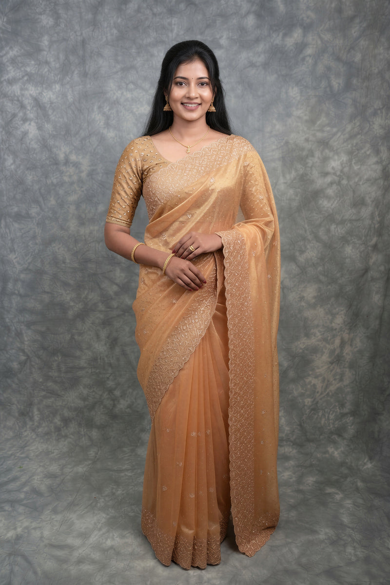 A woman modeling a peach-beige embroidered saree with a matching embroidered blouse, standing against a mottled gray studio backdrop.
