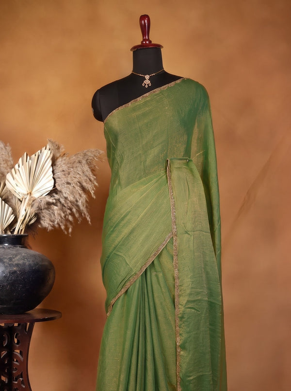 An olive green silk saree draped on a black mannequin, styled with a gold necklace, set against a warm brown studio backdrop with a decorative dark ceramic vase and dried palm leaves on a wooden stand.