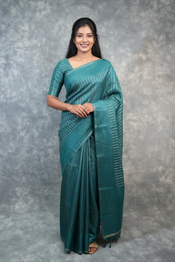 A woman modeling a teal green silk saree with vertical metallic gold stripes, standing against a textured gray studio backdrop.