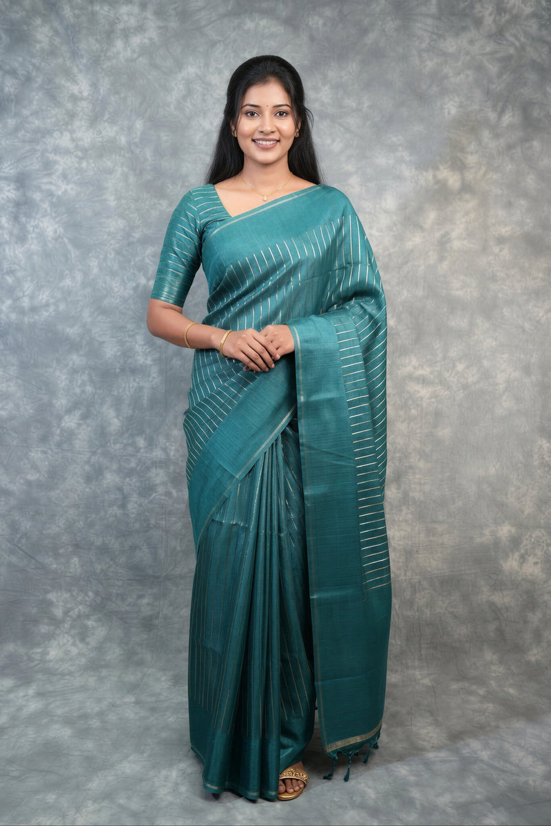 A woman modeling a teal green silk saree with vertical metallic gold stripes, standing against a textured gray studio backdrop.