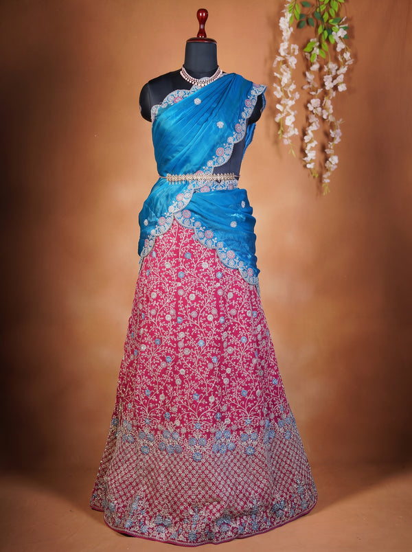 A traditional Indian lehenga choli set displayed on a black mannequin. The choli is bright blue with silver embroidery and a matching waist belt, while the long skirt is pink with intricate floral embroidery and a blue patterned border.