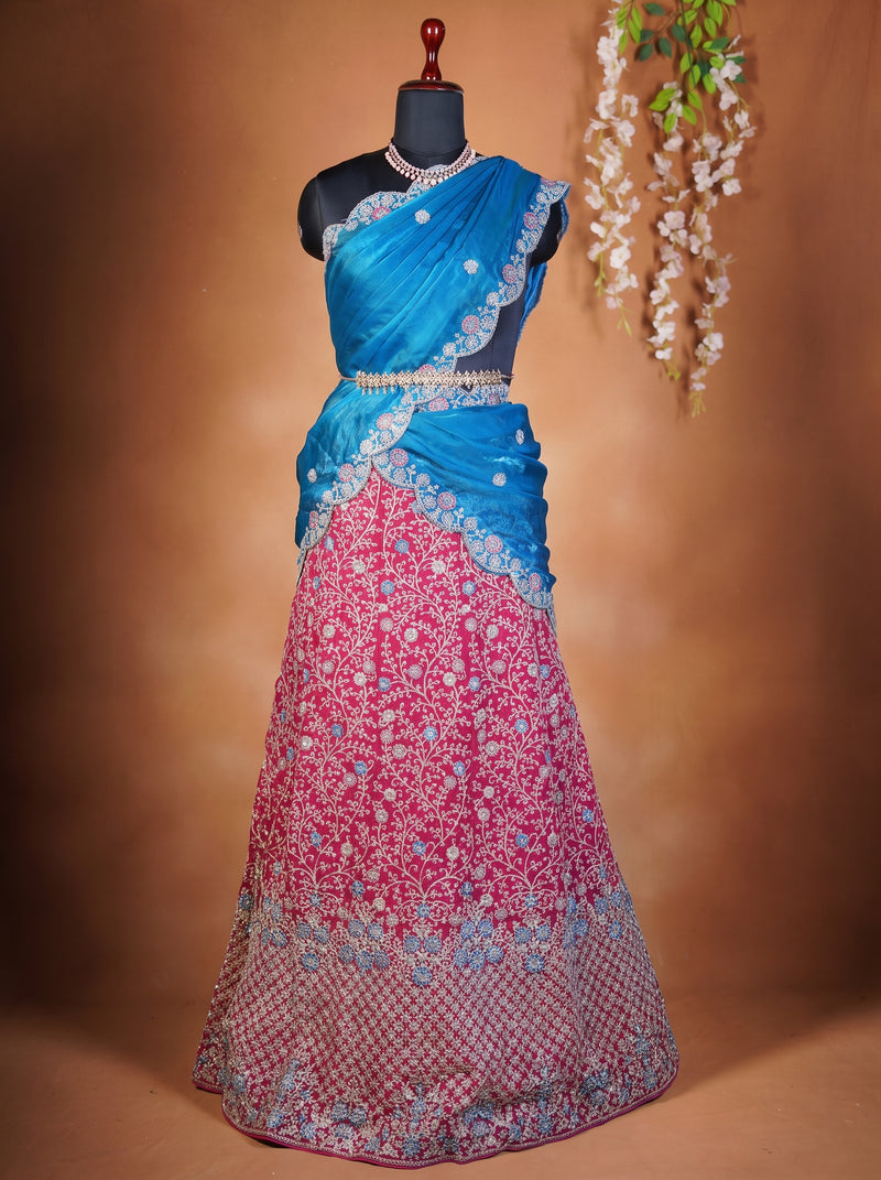 A traditional Indian lehenga choli set displayed on a black mannequin. The choli is bright blue with silver embroidery and a matching waist belt, while the long skirt is pink with intricate floral embroidery and a blue patterned border.