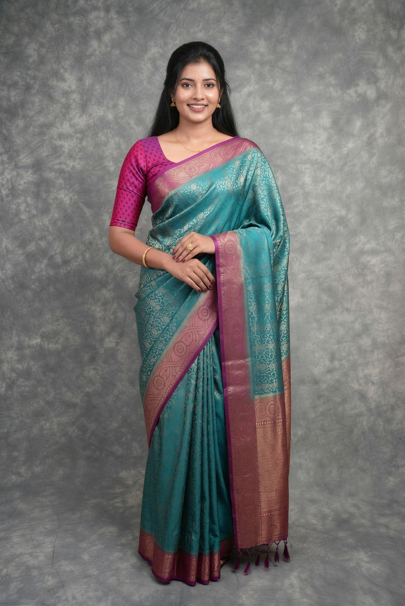 A woman modeling a teal and magenta silk saree with a metallic bronze border, standing against a textured gray studio backdrop.