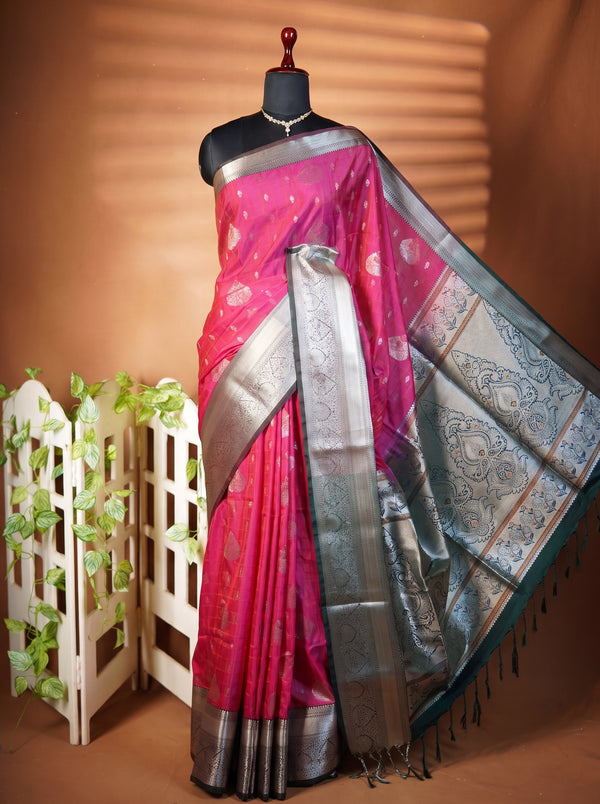 A pink and silver Banarasi silk saree displayed on a black mannequin torso, styled with a decorative white wooden screen and green artificial vines against a warm brown background.