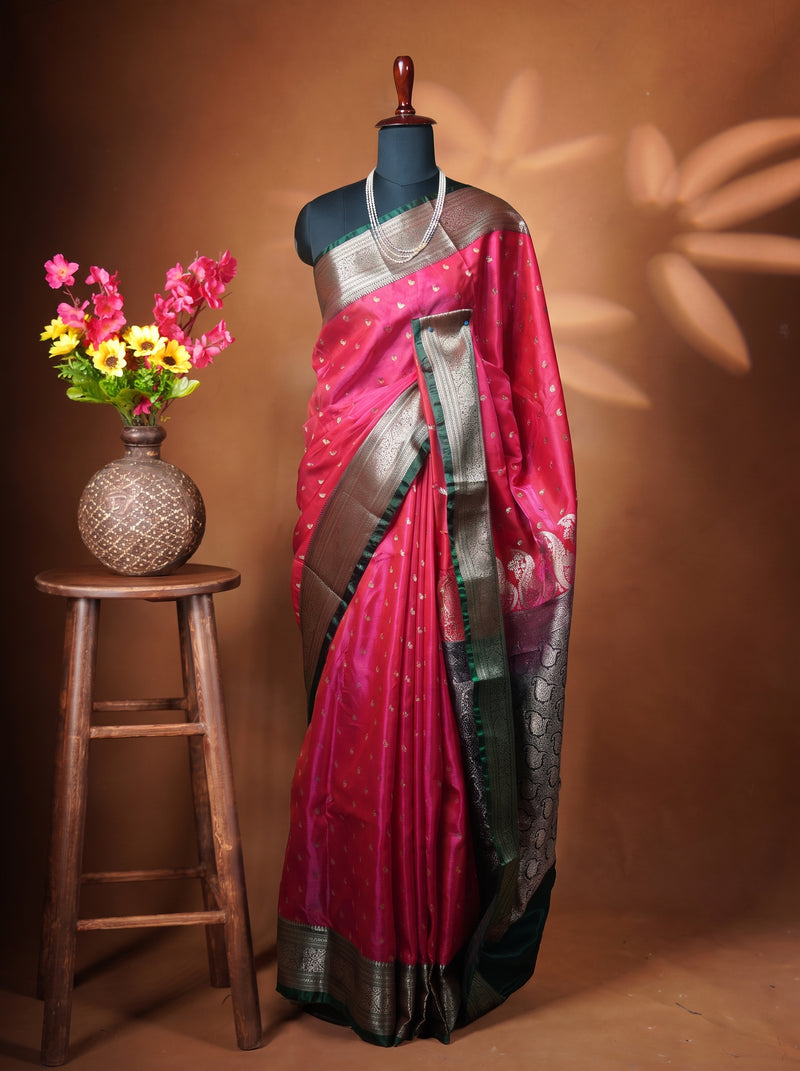 A magenta pink silk saree with metallic silver and gold zari borders, displayed on a black mannequin torso. The mannequin is styled with a silver necklace and stands next to a wooden stool holding a vase of pink and yellow flowers against a warm brown background with leaf shadows.