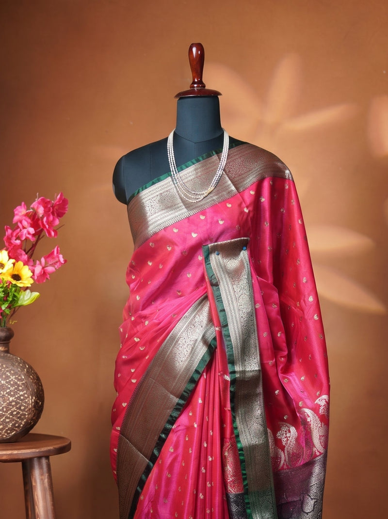 A magenta pink silk saree with metallic silver and gold zari borders, displayed on a black mannequin torso. The mannequin is styled with a silver necklace and stands next to a wooden stool holding a vase of pink and yellow flowers against a warm brown background with leaf shadows.
