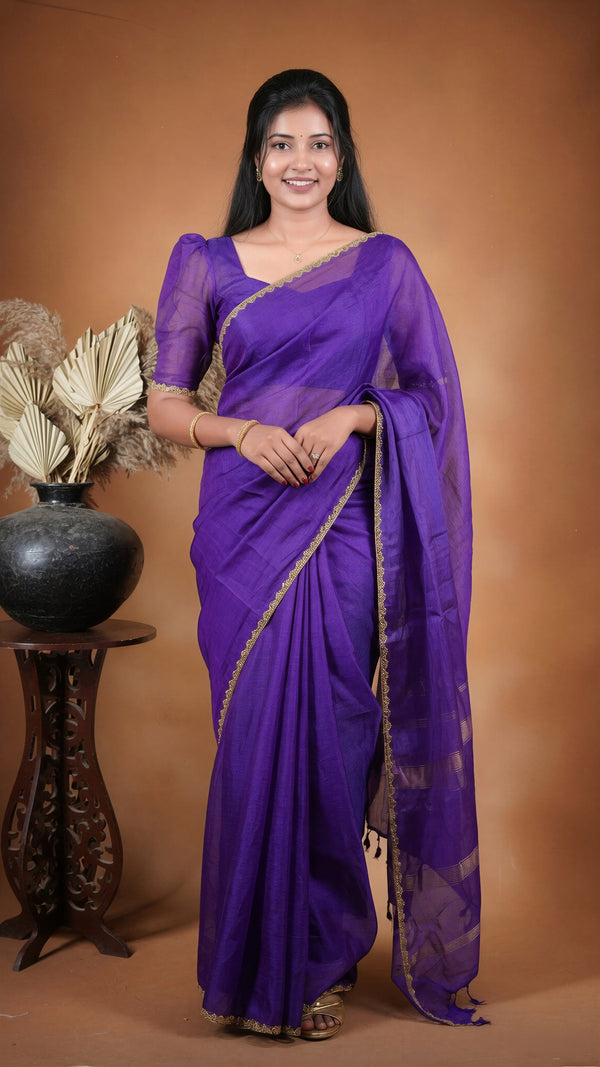 A woman modeling a purple silk saree with gold zari embroidery, standing against a warm brown studio backdrop next to a decorative vase.