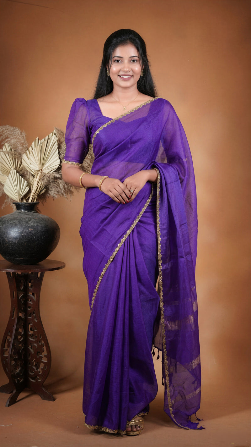A woman modeling a purple silk saree with gold zari embroidery, standing against a warm brown studio backdrop next to a decorative vase.