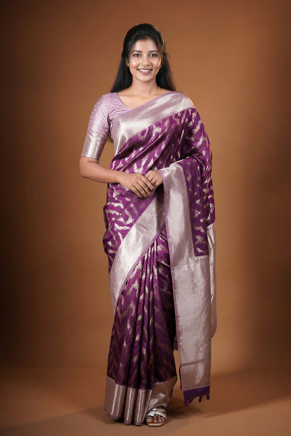A woman modeling a purple and silver patterned silk saree with a matching silver blouse, standing against a solid brown studio background.
