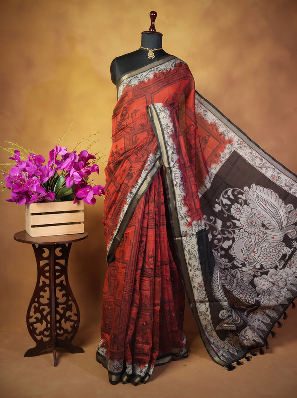 A red and black printed saree displayed on a black mannequin, with a large black and white patterned border draped over the shoulder. The mannequin stands against a warm brown studio backdrop next to a small table holding a box of purple flowers.