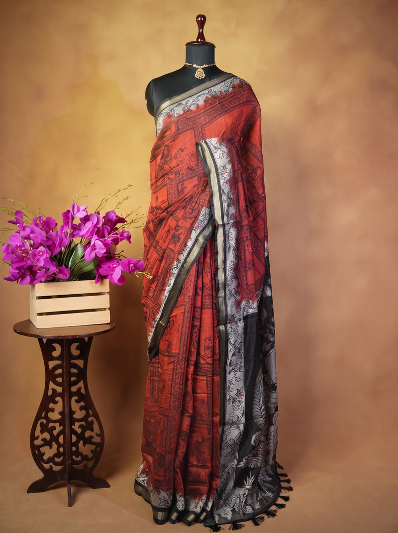 A printed saree displayed on a black dress form mannequin, featuring a rust red and black patterned body with a gray and black border. The mannequin is set against a warm brown studio backdrop, with a small wooden stand holding a vase of purple flowers on the left.