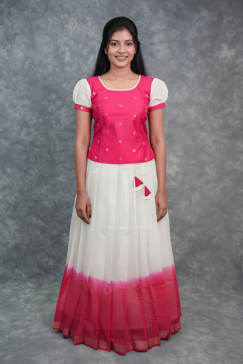 A young girl modeling a traditional Indian dress consisting of a bright pink short-sleeve top with gold embroidery and a white tiered skirt with a pink ombre hem, set against a gray textured studio backdrop.