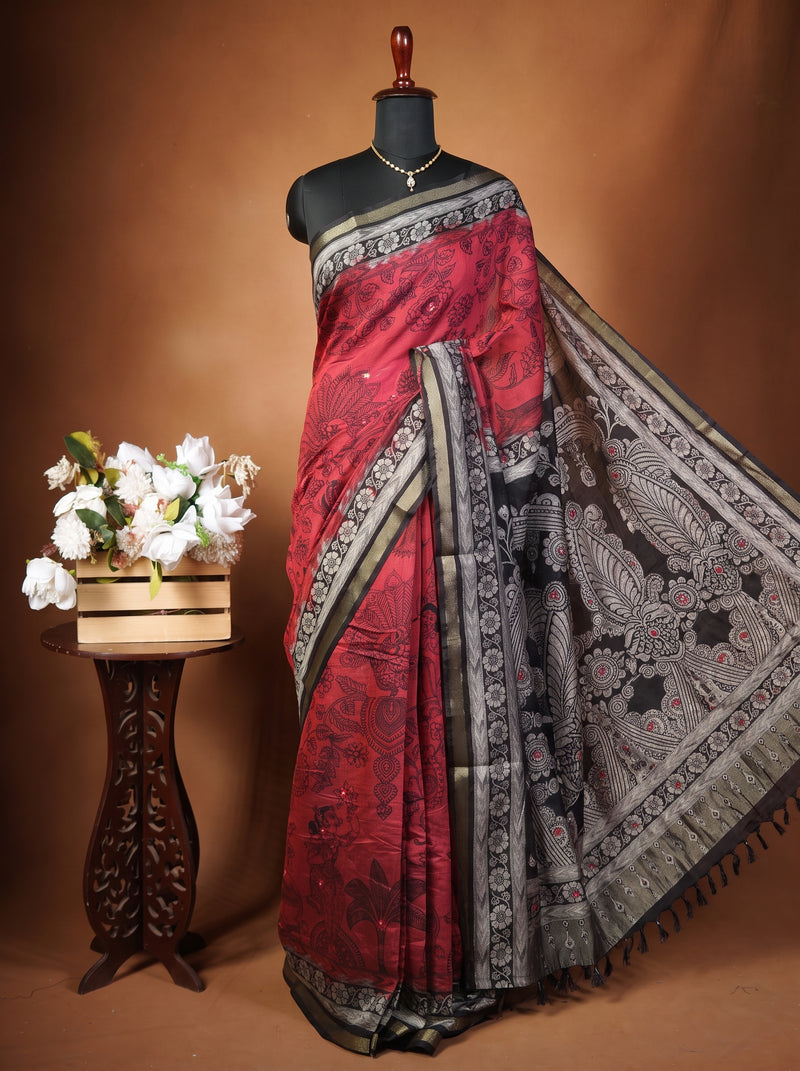 A red and black silk saree with intricate silver and gold borders, displayed on a black mannequin torso. The mannequin is set against a warm brown studio backdrop, with a decorative wooden stand holding white flowers on the left.