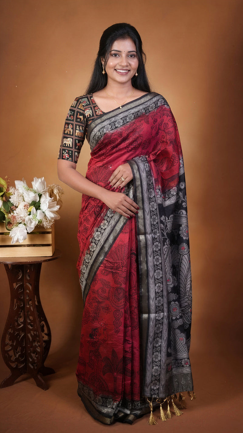 A woman modeling a red and black patterned saree with a black patterned blouse, standing against a warm brown studio backdrop next to a decorative table with white flowers.