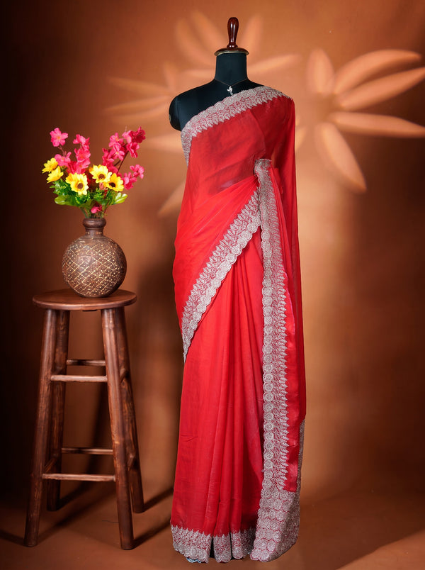 A red saree with a wide silver embroidered border is draped on a black mannequin torso. The mannequin is set against a warm brown background with decorative leaf shadows, and a small wooden stool holding a vase of colorful flowers is on the left.